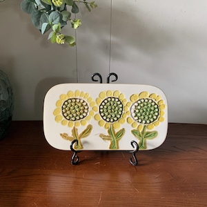May include: A rectangular ceramic tray featuring a raised, textured design of three sunflowers with yellow petals and green centers. The tray has a white background and is displayed on a black metal stand, showcasing a relief pattern of stems.