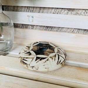 May include: A round, off-white decorative stone with intricate carved floral patterns. The center features a dark, marbled design. The stone is placed on a light-colored wooden surface, suggesting a decorative or artistic item.