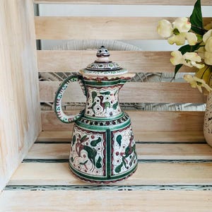 May include: A ceramic pitcher with a lid, featuring hand-painted green and brown illustrations of deer and birds. The vessel has a wide base, a tapered neck, a curved handle, and a pointed spout, all decorated with intricate geometric border patterns.