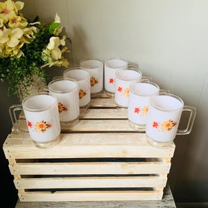 May include: A set of eight white glass mugs with clear handles. Each mug features a floral design in shades of yellow, orange, and red. The mugs are arranged on a wooden crate, with a bouquet of yellow flowers in the background.