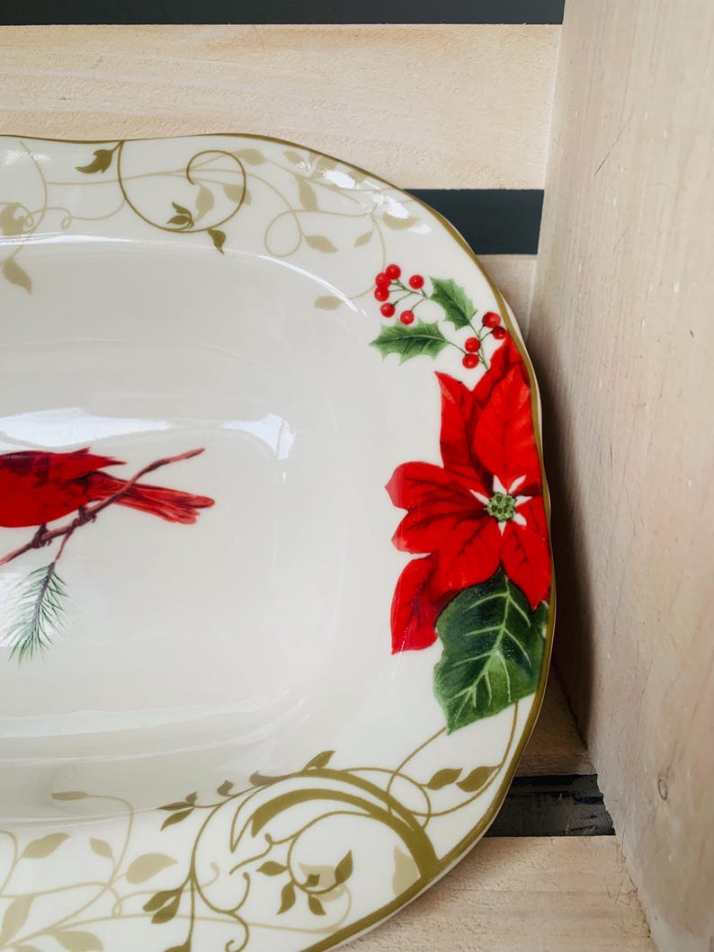 May include: White oval serving dish with a gold rim and a Christmas design. The design features a red cardinal bird, red poinsettia flowers, and green leaves.