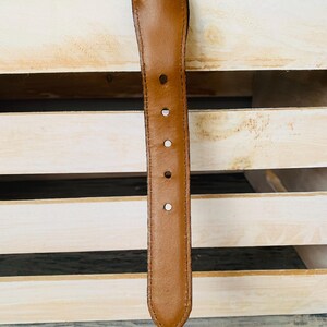 Puede incluir: Primer plano de un cintur&oacute;n de cuero marr&oacute;n con cinco agujeros y un extremo redondeado. El cintur&oacute;n est&aacute; colocado contra una caja de madera de color claro, con una correa tejida visible en el fondo. Un accesorio cl&aacute;sico.