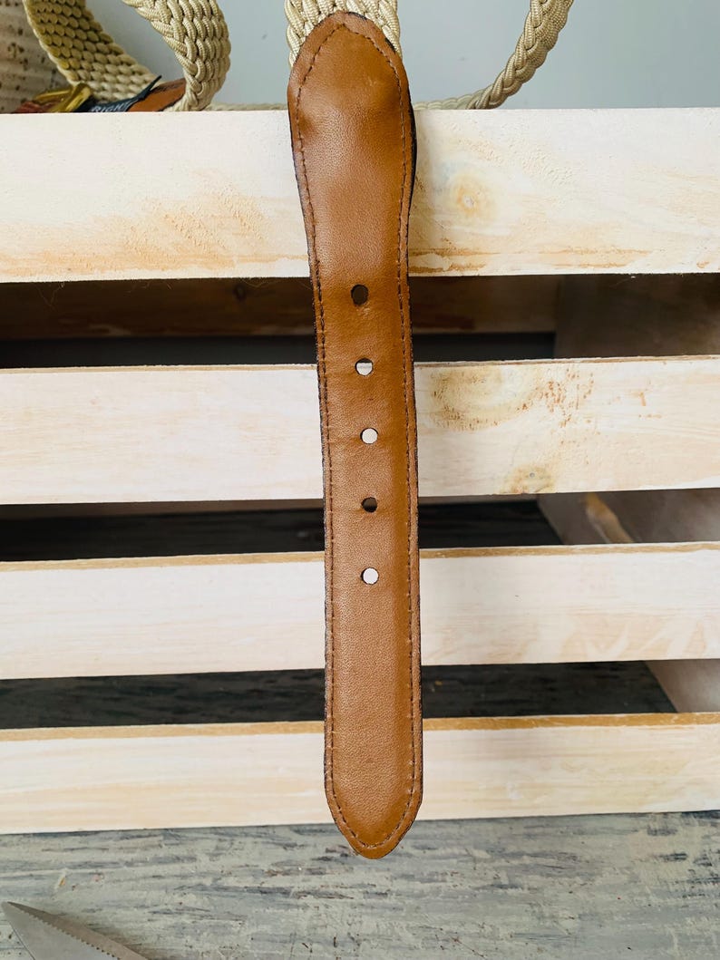 Puede incluir: Primer plano de un cintur&oacute;n de cuero marr&oacute;n con cinco agujeros y un extremo redondeado. El cintur&oacute;n est&aacute; colocado contra una caja de madera de color claro, con una correa tejida visible en el fondo. Un accesorio cl&aacute;sico.