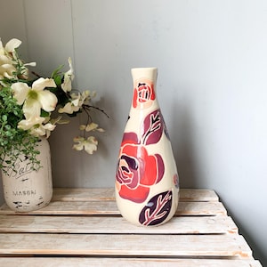 May include: Cream-colored ceramic vase with a floral design in shades of red and purple. The vase features a slender neck and a rounded body. The flowers and leaves are painted on the vase. The vase is displayed on a wooden crate.