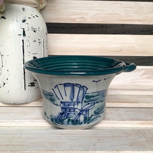 May include: A ceramic bowl with a teal rim and handle. The bowl features a blue and white beach scene with a chair. The bowl is on a wooden surface.