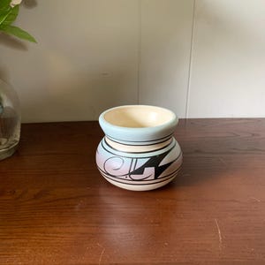 May include: A small, rounded ceramic pot with a cream-colored interior and a light blue rim. The exterior features a gradient of soft pink and blue hues, accented by bold, black geometric lines and abstract shapes encircling the body.