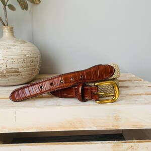 Puede incluir: Un cintur&oacute;n de cuero marr&oacute;n con una hebilla dorada y una secci&oacute;n de paja tejida. El cintur&oacute;n est&aacute; sobre una superficie de madera clara. La parte de cuero tiene un patr&oacute;n texturizado similar al de un cocodrilo.