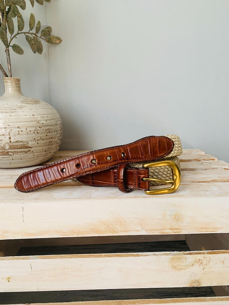 Puede incluir: Un cintur&oacute;n de cuero marr&oacute;n con una hebilla dorada y una secci&oacute;n de paja tejida. El cintur&oacute;n est&aacute; sobre una superficie de madera clara. La parte de cuero tiene un patr&oacute;n texturizado similar al de un cocodrilo.