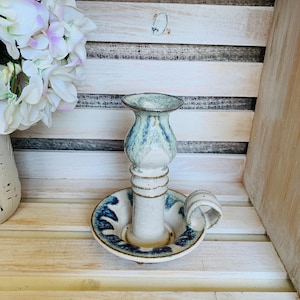 May include: A handmade ceramic candle holder with a white and blue glaze. The candle holder has a circular base with a handle and a detailed design. The candlestick is approximately 13 cm tall.