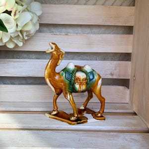 May include: A ceramic camel figurine with a brown and tan glaze, accented with green and blue details on its saddle. The camel is standing on a rectangular base. Decorative item.