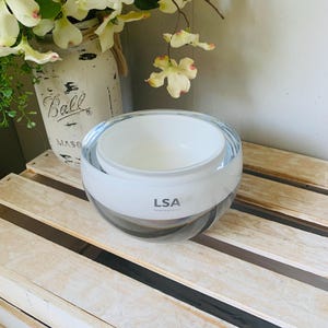 May include: A clear and white glass bowl with the text "LSA International" on the side. The bowl is sitting on a wooden surface. A white mason jar with flowers is in the background.