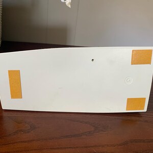 May include: A white, rectangular object with rounded edges, featuring four rectangular, tan-colored adhesive pads on the bottom. The object is resting on a dark brown wooden surface.