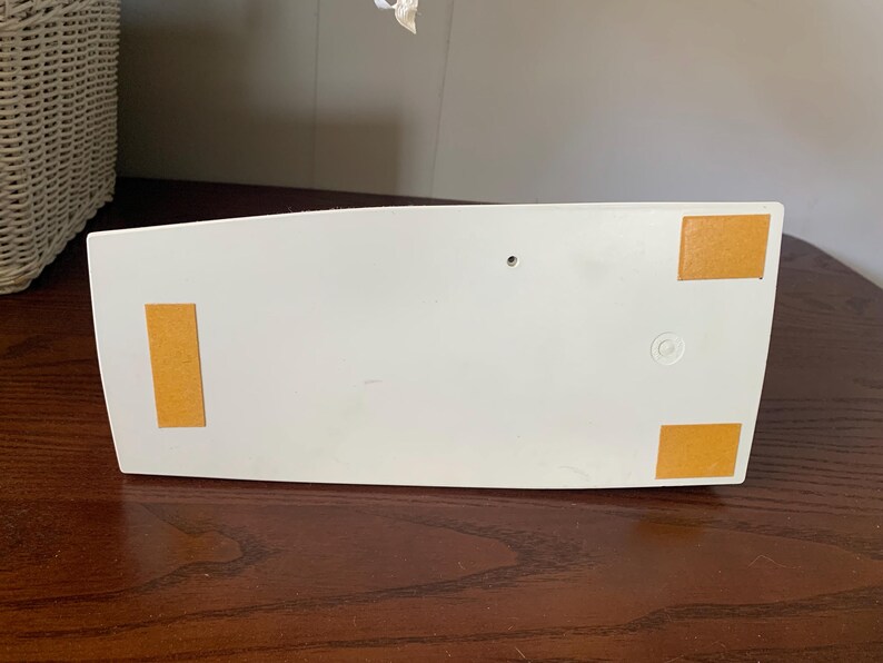 May include: A white, rectangular object with rounded edges, featuring four rectangular, tan-colored adhesive pads on the bottom. The object is resting on a dark brown wooden surface.