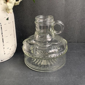 May include: Clear glass oil lamp with a handle. The lamp has a round base and a decorative, scalloped design around the middle. The top portion has a handle. The lamp is sitting on a dark surface.