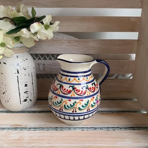 May include: A ceramic pitcher with a cream-colored base featuring hand-painted horizontal bands of blue, red, and green scrollwork patterns. The pitcher has a rounded body, a curved handle, and a flared spout, showcasing a glossy finish.