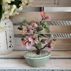 May include: A decorative bonsai tree sculpture featuring translucent pink flower petals with yellow centers and green leaves attached to dark brown branches. The tree is set in a small, light green ceramic pot filled with white gravel stones.