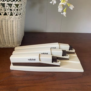 May include: A set of three vintage Wiltshire Stay Sharp staplers in a cream color with dark brown accents. The staplers are arranged on a matching cream-colored base, with the brand name printed on the side. The background includes a white wicker basket and floral arrangement.