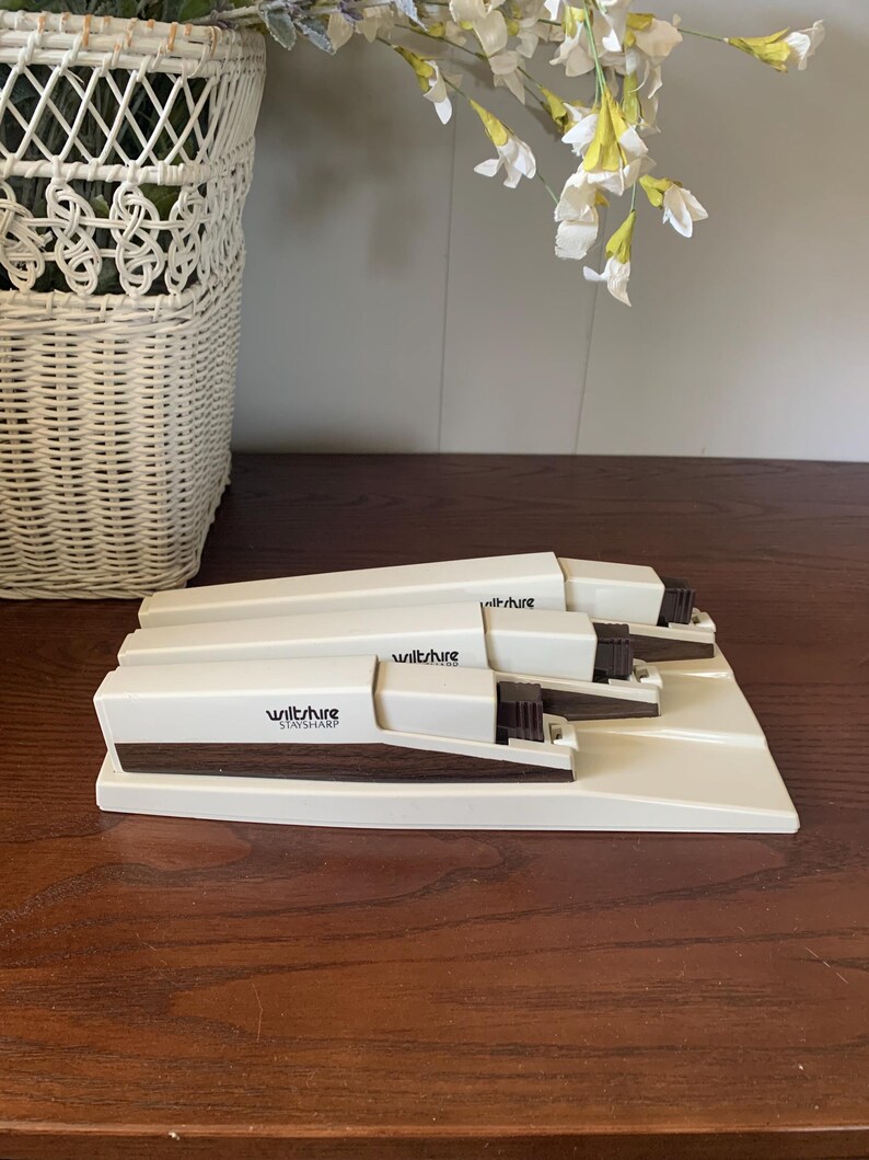 May include: A set of three vintage Wiltshire Stay Sharp staplers in a cream color with dark brown accents. The staplers are arranged on a matching cream-colored base, with the brand name printed on the side. The background includes a white wicker basket and floral arrangement.