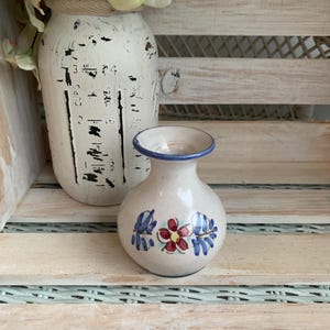 May include: A small, round, cream-colored ceramic vase with a narrow neck and a blue rim. The body of the vase features a hand-painted design of a central red flower with yellow center, flanked by blue leaves on either side of the floral motif.