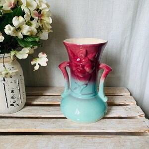 May include: A vintage ceramic vase with a two-tone design. The top is a deep burgundy, fading into a turquoise base. It features two curved handles and a floral relief pattern. The vase is displayed on a rustic wooden surface.