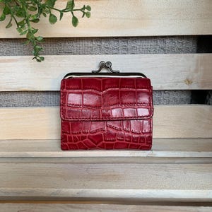 May include: A red, textured wallet with a kiss-lock clasp. The wallet has a crocodile skin pattern and a metal clasp. The wallet is sitting on a wooden surface.