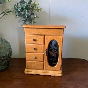 May include: A light brown wooden jewelry armoire featuring a top compartment, four small drawers with dark circular knobs, and a side door with an oval glass panel etched with a floral design. The cabinet has a rectangular base and top.