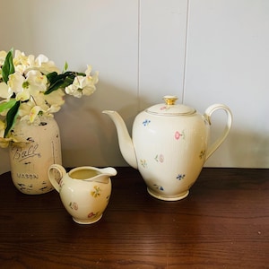 May include: A cream-colored porcelain tea set with a teapot and creamer, adorned with delicate floral patterns in pink, blue, and yellow. The teapot features a gold-colored handle and lid finial. A Ball Mason jar with white flowers is in the background.