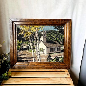 May include: Framed tapestry art depicting a white church with a steeple, a covered bridge, and trees. The artwork is set in a wooden frame. The scene includes birch trees with yellow leaves, and a green hillside in the background.