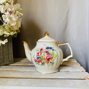May include: Cream-colored porcelain teapot with a floral basket design. The teapot features a gold-trimmed handle, spout, and lid. The floral basket includes pink roses, blue flowers, and green leaves. The teapot is set on a white wooden surface.