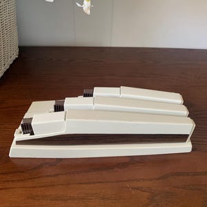 May include: A stack of off-white plastic blinds with dark brown accents. The blinds are rectangular and appear to be folded or stacked. The blinds are on a dark brown wooden surface.