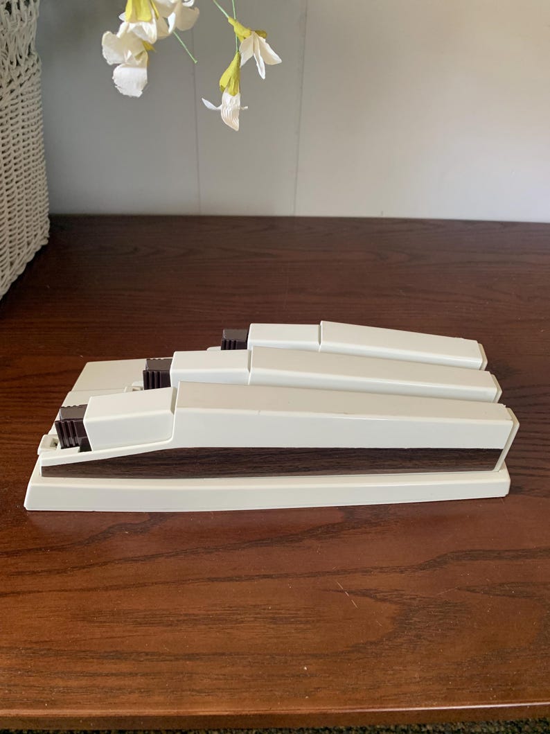 May include: A stack of off-white plastic blinds with dark brown accents. The blinds are rectangular and appear to be folded or stacked. The blinds are on a dark brown wooden surface.