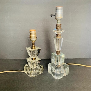 May include: Two clear glass table lamps with geometric designs. The lamps have a square base and a stacked, faceted body. Each lamp has a metal lamp holder and a power cord. The lamps are on a dark surface.