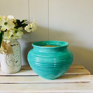 May include: A turquoise ceramic bowl with a rounded, bulbous shape and horizontal ribbed texture throughout the exterior. The bowl features a wide, open mouth and a smooth, glossy finish, showcasing a vibrant teal colour that contrasts with the surface.
