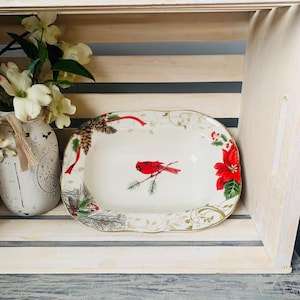 May include: A white oval serving dish with a gold rim and a festive Christmas design. The design features a red cardinal bird on a branch, pine cones, holly, and poinsettia flowers.