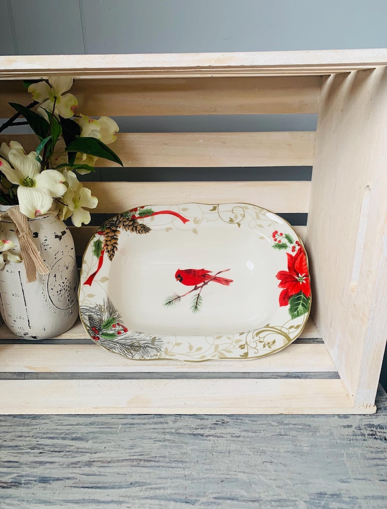 May include: A white oval serving dish with a gold rim and a festive Christmas design. The design features a red cardinal bird on a branch, pine cones, holly, and poinsettia flowers.