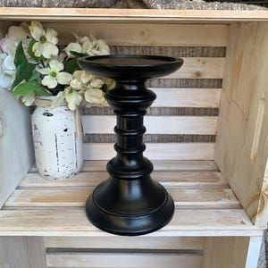 May include: A black, decorative candle holder with a wide, round top and a pedestal base. The holder is made of a solid material and has a matte finish. The candle holder is displayed on a white wooden shelf with a mason jar of flowers.