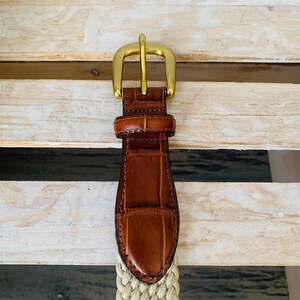 Puede incluir: Un cintur&oacute;n de cuero marr&oacute;n con una hebilla dorada. El cintur&oacute;n presenta una tela beige tejida y una punta de cuero marr&oacute;n con un patr&oacute;n de cocodrilo. La hebilla es de dise&ntilde;o cl&aacute;sico y sencillo.