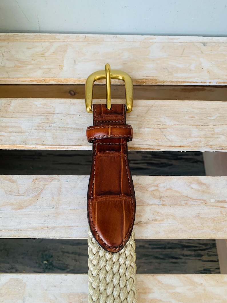 Puede incluir: Un cintur&oacute;n de cuero marr&oacute;n con una hebilla dorada. El cintur&oacute;n presenta una tela beige tejida y una punta de cuero marr&oacute;n con un patr&oacute;n de cocodrilo. La hebilla es de dise&ntilde;o cl&aacute;sico y sencillo.