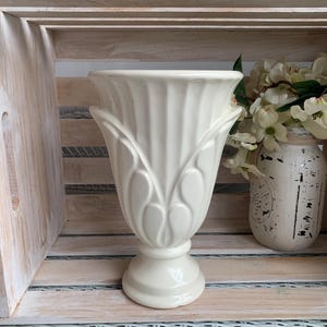 May include: A cream-coloured ceramic pedestal vase featuring a flared rim and vertical fluted detailing along the body. The vase has a smooth, glossy finish and a wide, circular base, standing upright with a symmetrical, urn-like silhouette.
