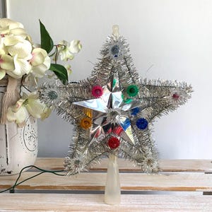 May include: A five-pointed star tree topper featuring a reflective silver metallic center and silver tinsel trim. The star is decorated with small, multi-colored light bulbs in red, blue, green, and yellow, and includes a clear plastic base.