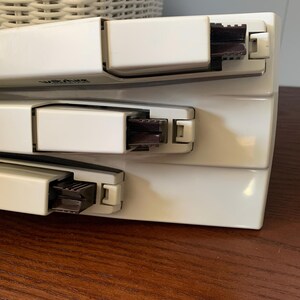 May include: Three white staplers stacked on top of each other. Each stapler has a dark brown stapling mechanism and a white lever. The word "Wiltshire" is printed on the top stapler. The staplers are on a wooden surface.