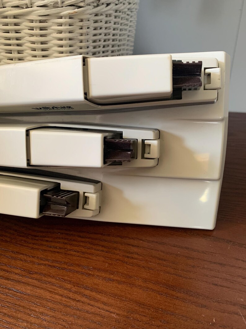 May include: Three white staplers stacked on top of each other. Each stapler has a dark brown stapling mechanism and a white lever. The word "Wiltshire" is printed on the top stapler. The staplers are on a wooden surface.