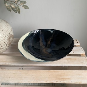 May include: A shallow ceramic bowl with a glossy finish, featuring a deep black interior with subtle blue and brown streaks. The exterior rim is accented with a wide, creamy off-white band that contrasts against the dark, reflective center.