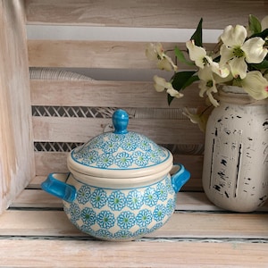 May include: A ceramic lidded tureen featuring a repeating blue and green floral pattern on a cream background. The round vessel has two side handles and a matching domed lid with a small blue knob handle at the top, showcasing a classic design.