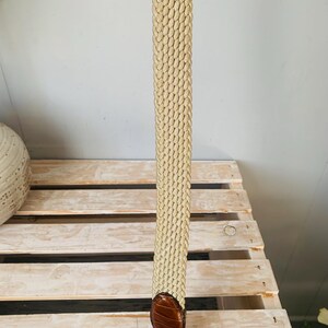 Puede incluir: Un cintur&oacute;n beige tejido con un extremo de cuero marr&oacute;n. El cintur&oacute;n tiene un dise&ntilde;o trenzado y un acabado de cuero texturizado. El cintur&oacute;n se muestra verticalmente sobre un fondo claro.