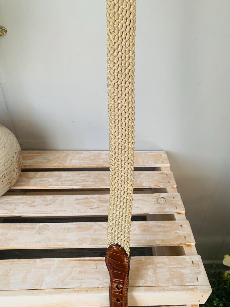 Puede incluir: Un cintur&oacute;n beige tejido con un extremo de cuero marr&oacute;n. El cintur&oacute;n tiene un dise&ntilde;o trenzado y un acabado de cuero texturizado. El cintur&oacute;n se muestra verticalmente sobre un fondo claro.