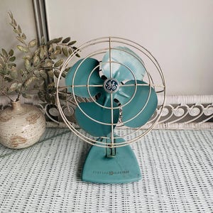 May include: A vintage General Electric desk fan featuring a teal metal base and four matching teal blades. The fan is protected by a circular white wire cage and includes a central hub with the GE logo. The base is marked General Electric.