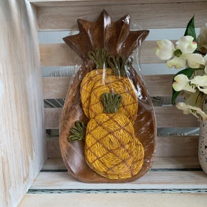 May include: A pineapple-shaped wooden bowl featuring a dark brown finish and a pointed top. Inside the bowl are two circular coasters made of yellow woven fiber with green stitched details, designed to resemble the texture of a pineapple.