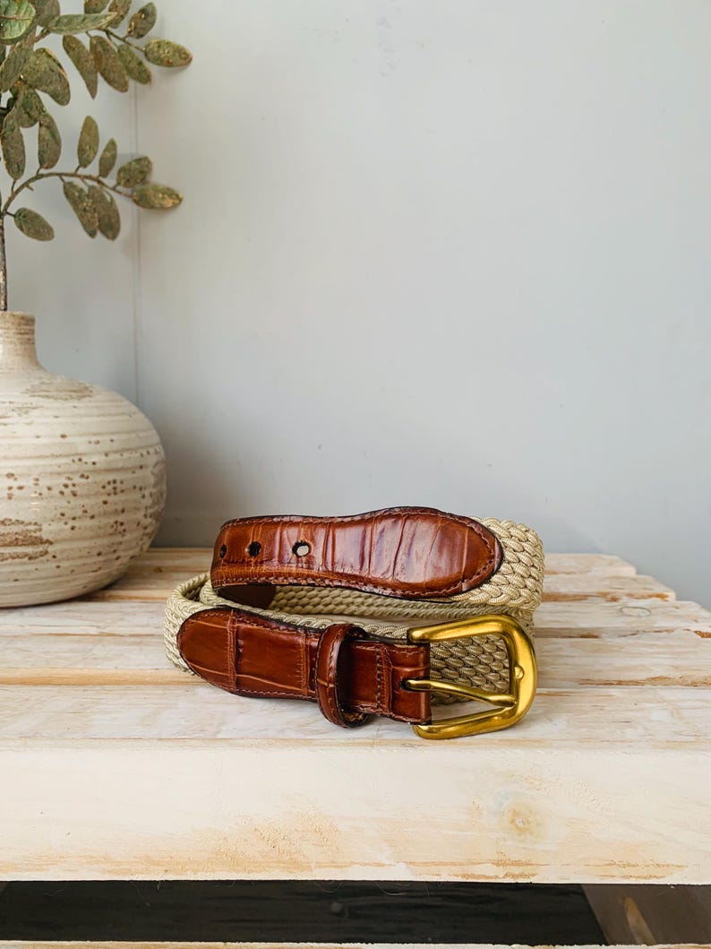 Puede incluir: Un cintur&oacute;n trenzado beige con detalles de cuero marr&oacute;n y una hebilla dorada. El cintur&oacute;n presenta un dise&ntilde;o texturizado y un estilo cl&aacute;sico. El cuero tiene un patr&oacute;n de cocodrilo.