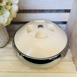 May include: Cream-colored, round container with a shiny chrome band. The top has four recessed areas and a small, rectangular emblem. The container sits on a light-colored wooden surface, suggesting a vintage or retro aesthetic.